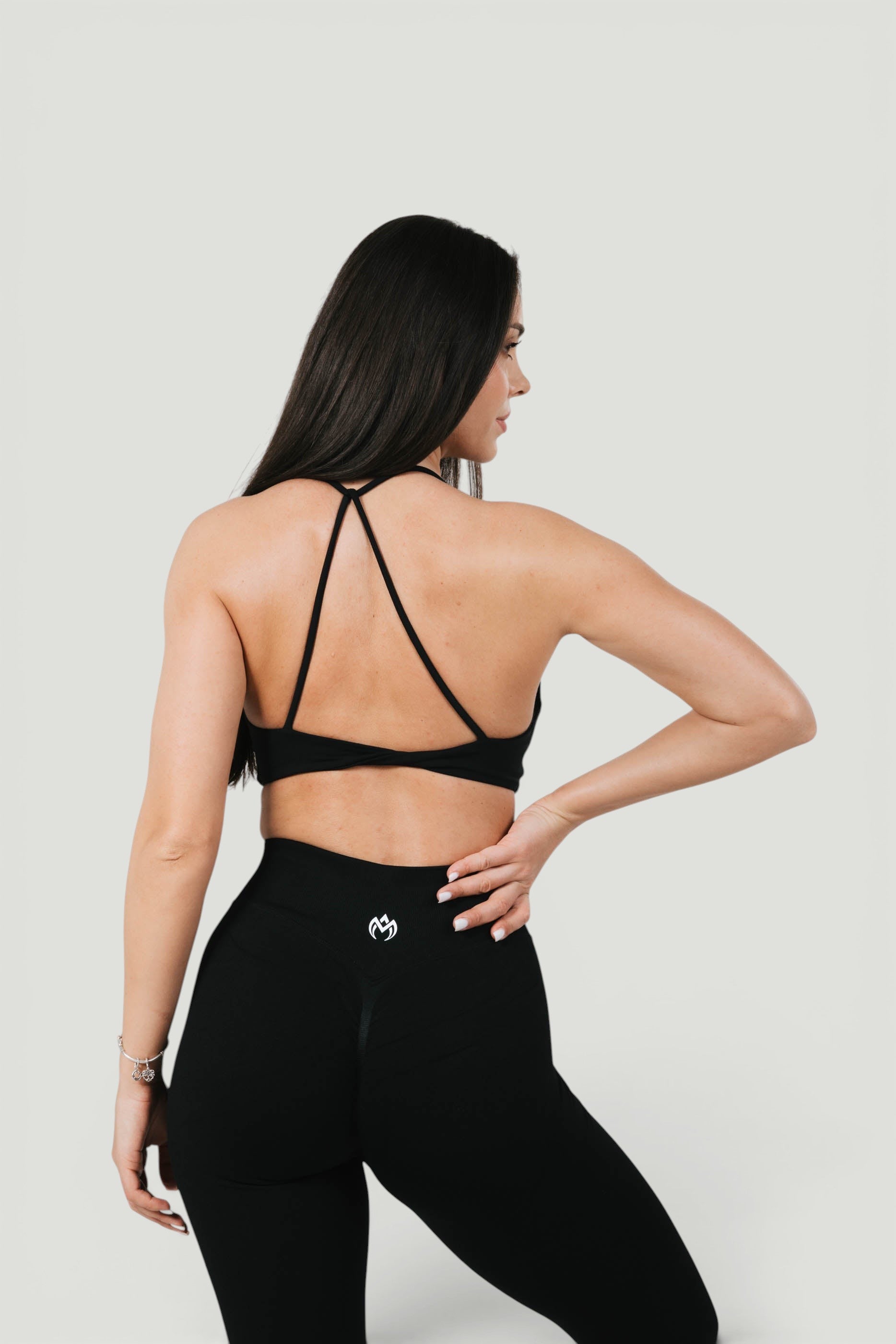 A N A M E black midnight sports bra with delicate straps and a twisted-back detail worn by a woman against a light background.